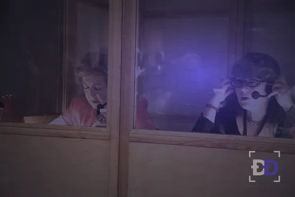 Two interpreters in glass booths wear headsets with microphones while they translate a meeting.