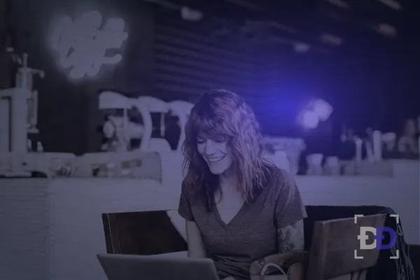 A woman smiles at her laptop while sitting at a table in a coffee shop.